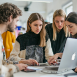 Quattro persone sedute intorno a un tavolo, concentrate su un computer portatile, collaborano e discutono di idee con una mentalità imprenditoriale in un ambiente d'ufficio luminoso e moderno.