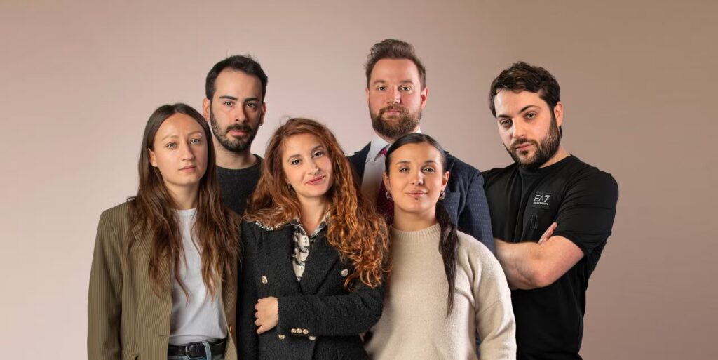 Sei adulti si trovano insieme su uno sfondo semplice, in posa per una foto di gruppo in abiti casual e business casual: l'ispirazione ideale per Produzione Abbigliamento Conto Terzi Piccole Quantità. La maggior parte ha un'espressione neutra; due sorridono.