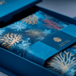 Una confezione regalo blu contenente una stoffa piegata a tema oceanico con motivi di corallo oro, bianco e rosso - ispirazione perfetta se volete creare un marchio di costumi da bagno - fissata da un nastro blu con scritte dorate e un disegno di corallo.